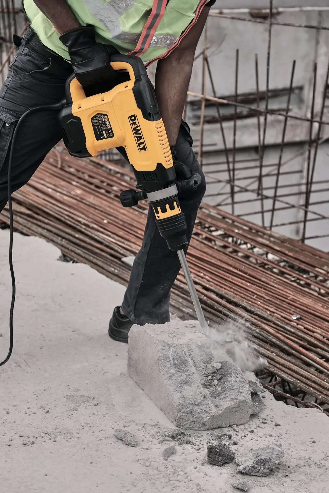 Una persona con ropa de trabajo utiliza un martillo demoledor amarillo DEWALT para romper un bloque de concreto en una obra de construcción.