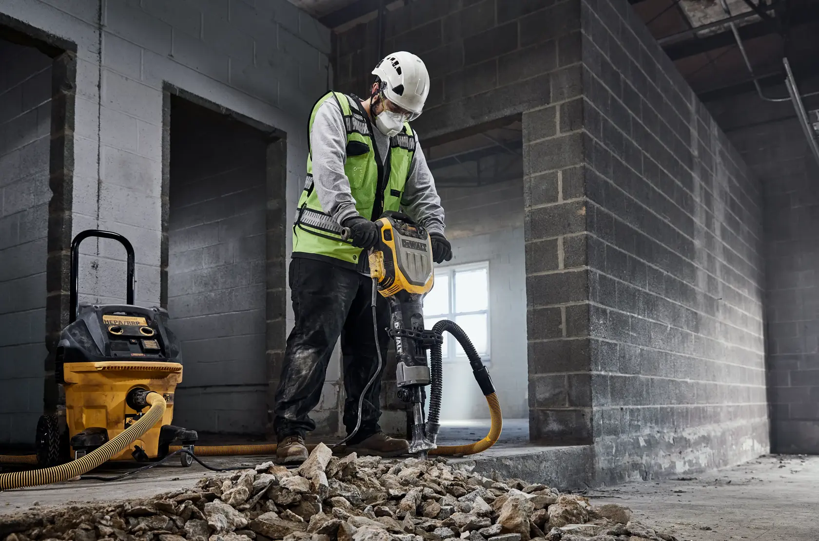 Un trabajador de la construcción con chaleco de seguridad y casco utiliza un martillo demoledor DEWALT (modelo D25966) para romper concreto en el interior de un edificio en construcción. Un extractor de polvo DEWALT está conectado al martillo demoledor y escombros de concreto roto se esparcen por el suelo.