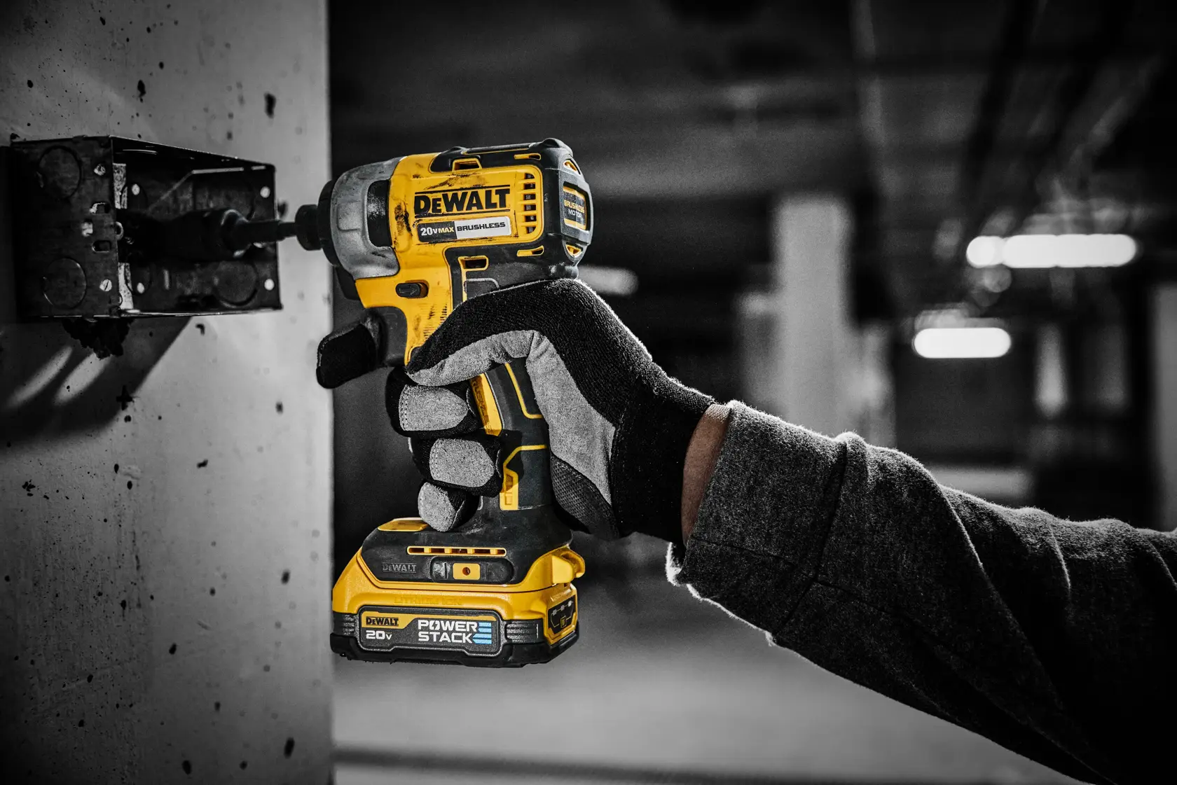 Una persona con guantes de trabajo usa un atornillador de impacto inalámbrico DEWALT de 20V con batería POWERSTACK para ensamblar o desmontar una caja eléctrica de metal montada en una pared de concreto en un entorno industrial.