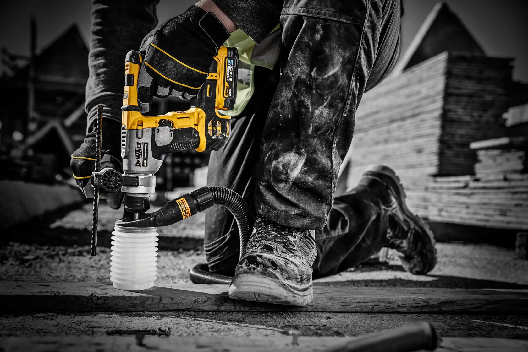 Un trabajador usa una herramienta eléctrica inalámbrica amarilla DEWALT con un accesorio de aspiradora para perforar una superficie en un sitio de construcción. Solo se ven las manos enguantadas, las piernas y el calzado del trabajador.
