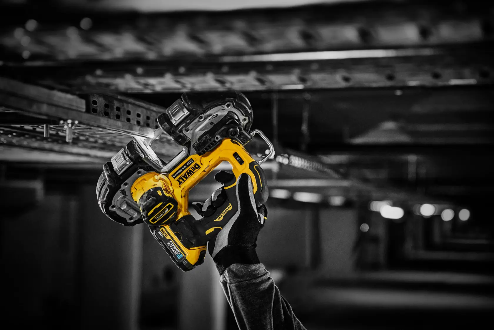 Una mano enguantada sostiene una herramienta eléctrica inalámbrica amarilla y negra DEWALT, trabajando sobre una estructura metálica en un entorno industrial con poca luz.
