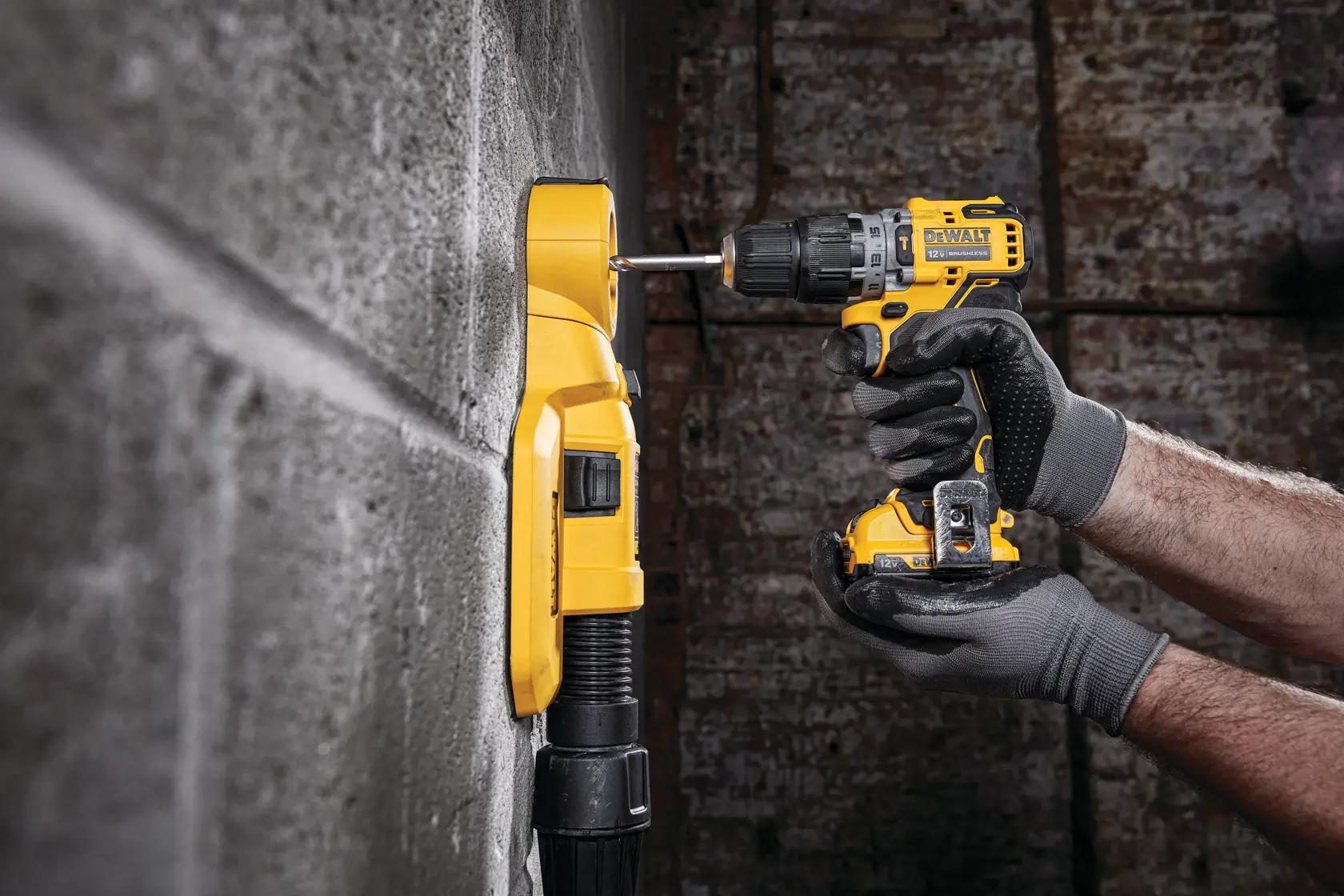 Una persona con guantes de trabajo grises está usando un taladro inalámbrico amarillo DEWALT para perforar una pared de concreto. Un dispositivo amarillo de extracción de polvo está colocado en la pared para recoger los residuos durante la perforación.