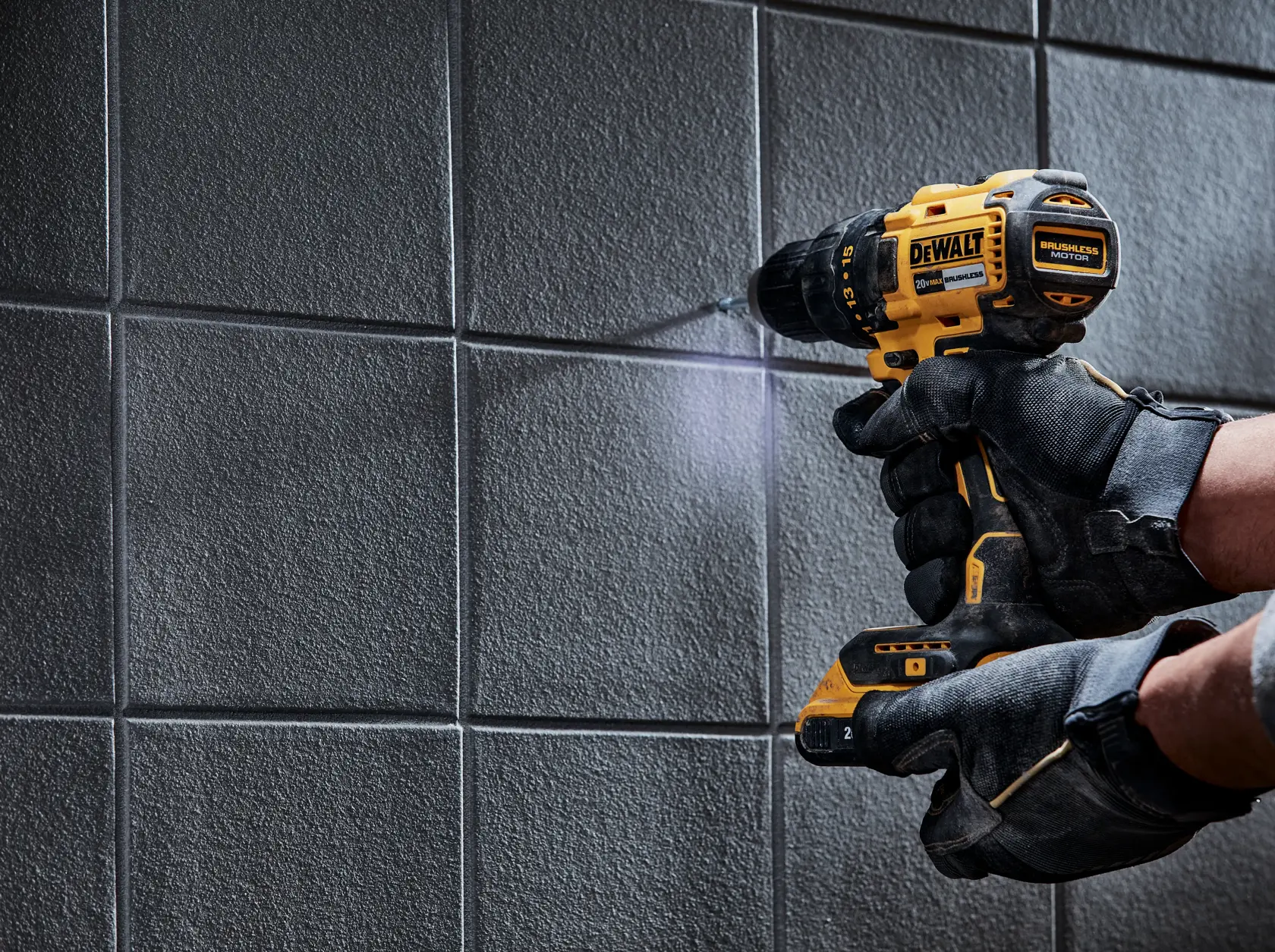 Una persona con guantes de trabajo negros está usando un taladro inalámbrico amarillo y negro DEWALT para perforar una pared de azulejos.