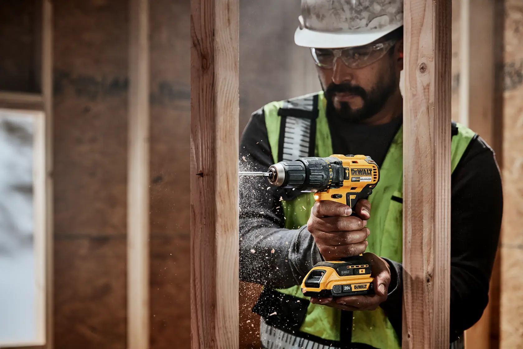 Un trabajador de la construcción con chaleco de seguridad y casco utiliza un taladro inalámbrico amarillo DEWALT para perforar una viga de madera, con partículas de madera volando desde el lugar de perforación.