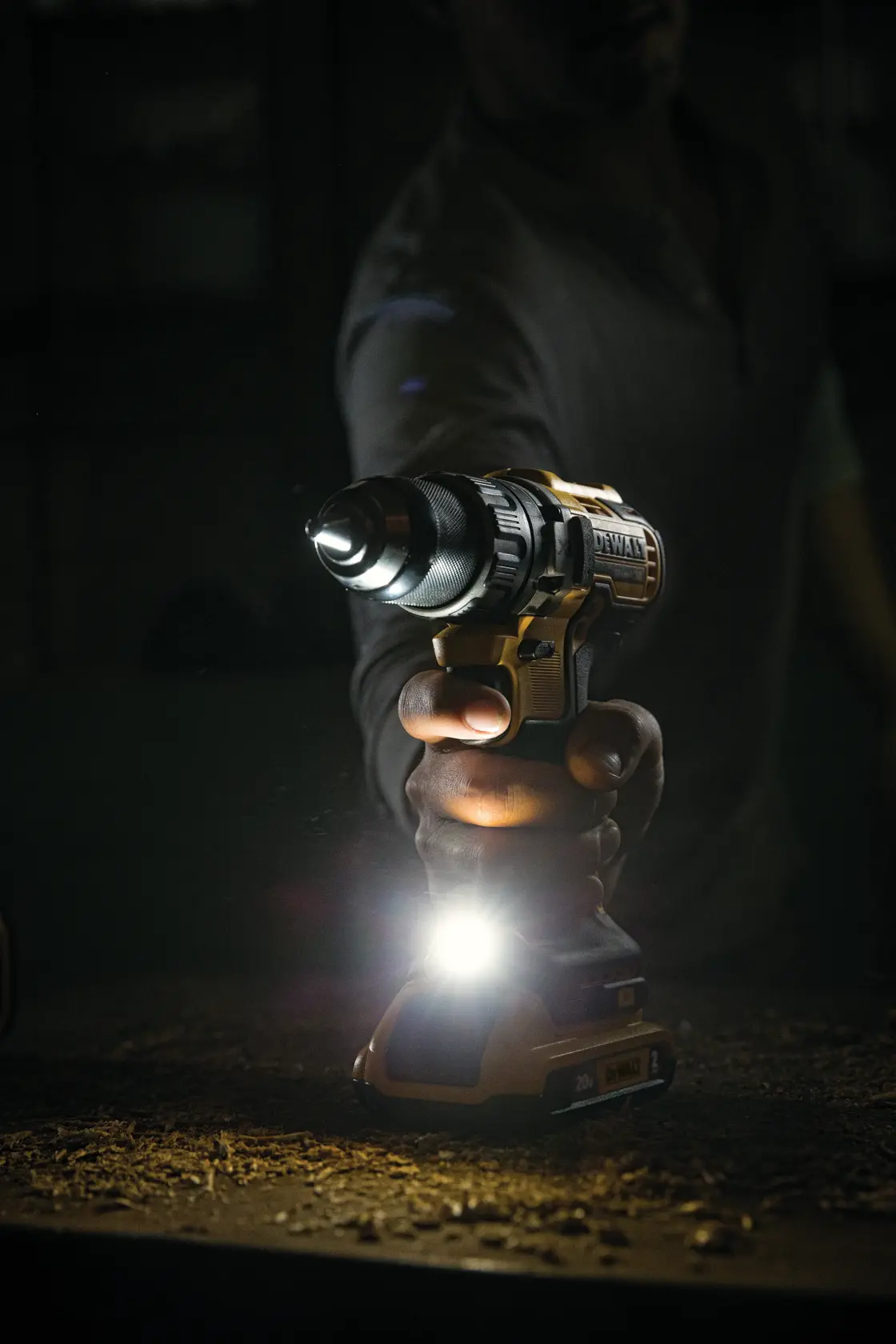 Una persona sostiene un taladro inalámbrico DEWALT con luz LED integrada encendida, iluminando la superficie de trabajo en un entorno con poca luz.
