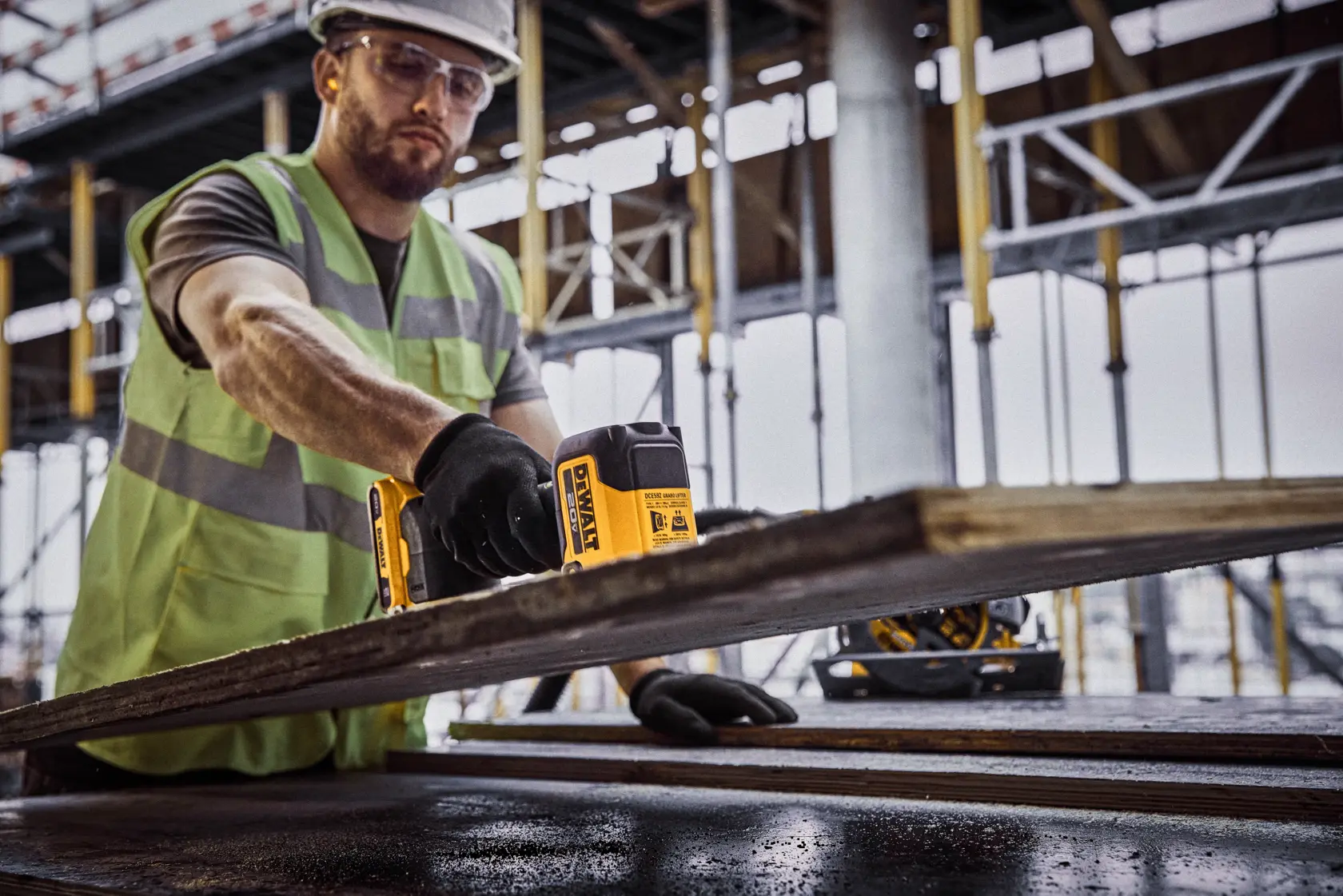 Una persona con chaleco de seguridad y guantes utiliza una herramienta DEWALT para medir o inspeccionar una tabla de madera en un sitio de construcción.