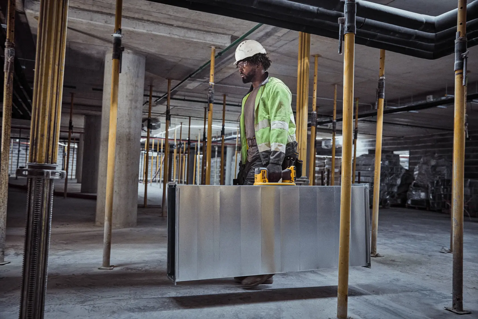 Un trabajador de la construcción con chaqueta de alta visibilidad y casco lleva un gran conducto metálico con la ayuda de una herramienta eléctrica amarilla DEWALT dentro de un edificio en construcción.