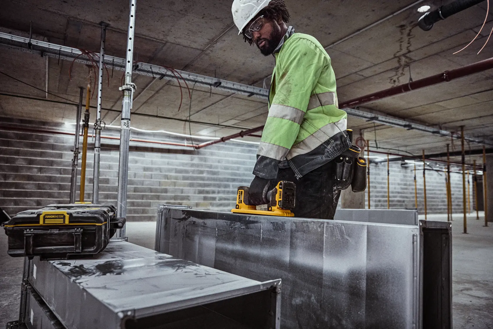 Un trabajador de la construcción con chaqueta de alta visibilidad utiliza una cámara de inspección inalámbrica DEWALT (DCE592B) para examinar un conducto metálico dentro de un edificio en construcción. Un estuche protector se encuentra cerca.