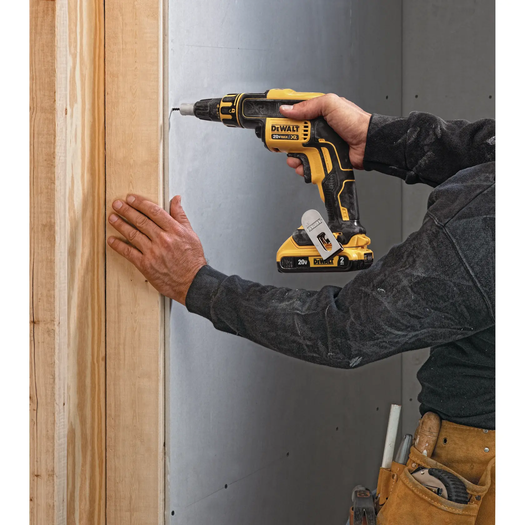 Una persona utiliza un atornillador inalámbrico DEWALT 20V MAX XR para fijar una placa de yeso a un marco de madera. La herramienta es amarilla y negra, y la persona lleva un cinturón de herramientas y ropa de trabajo.
