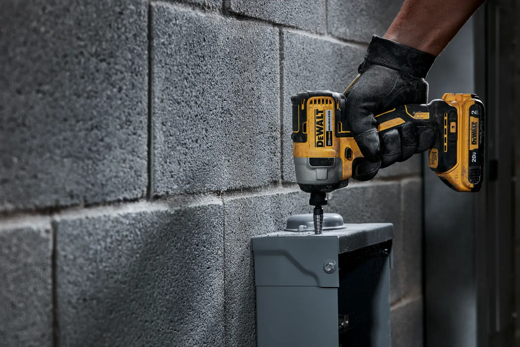 Una persona con guantes negros utiliza un destornillador de impacto inalámbrico DEWALT amarillo y negro para atornillar un tornillo en una caja eléctrica metálica montada en una pared de concreto.