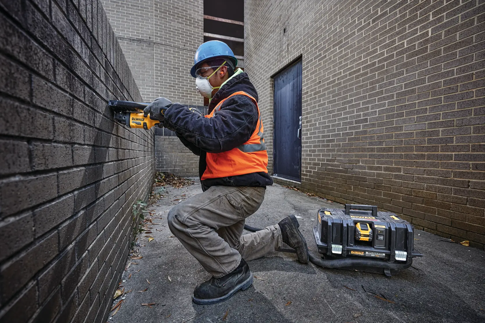 Un trabajador de la construcción con casco azul, chaleco de seguridad naranja, guantes y botas de trabajo está arrodillado usando una herramienta eléctrica amarilla DEWALT contra una pared de ladrillo. Junto al trabajador, en el suelo, hay una caja de herramientas portátil DEWALT. El rostro del trabajador está desenfocado.