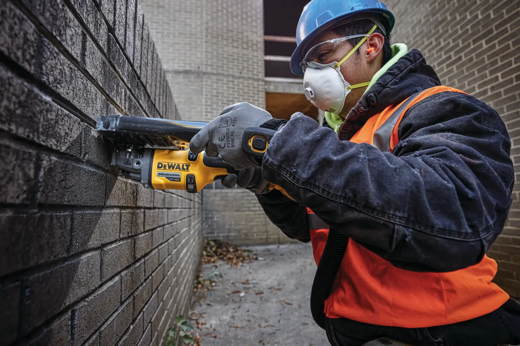 Una persona con casco azul, guantes y chaleco de seguridad naranja está usando una herramienta eléctrica inalámbrica DEWALT para cortar una pared de ladrillo. La herramienta tiene la marca DEWALT y parece estar en uso en un sitio de construcción al aire libre.