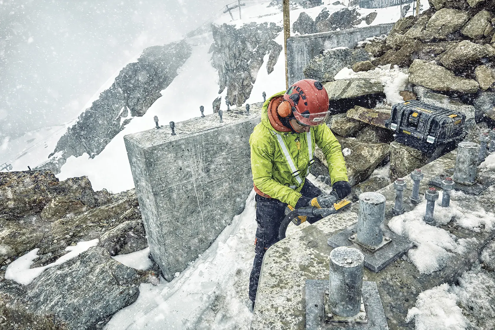 Una persona con equipo de seguridad utiliza una herramienta eléctrica DEWALT para trabajar en una estructura metálica al aire libre, en condiciones montañosas y nevadas. Un maletín negro de herramientas DEWALT descansa sobre una roca cercana.
