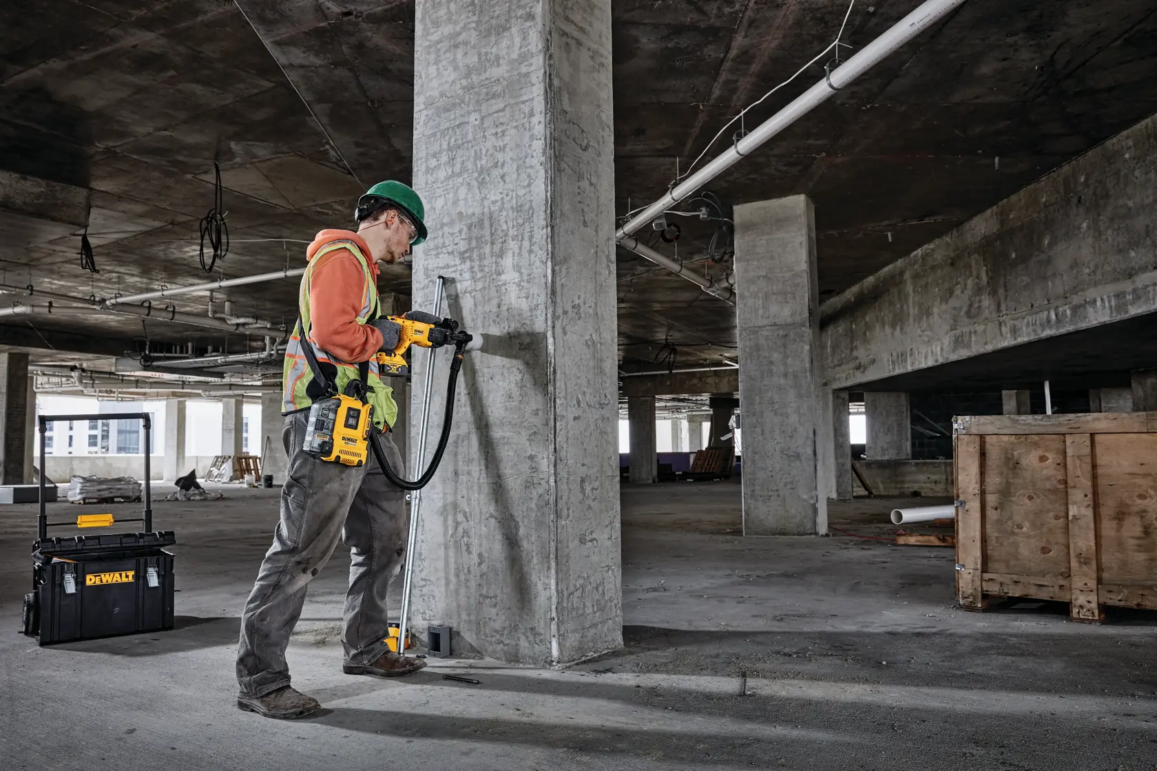 Un trabajador de la construcción utiliza un taladro rotativo DEWALT para perforar un pilar de concreto dentro de un edificio parcialmente construido. Cerca, hay un estuche de herramientas DEWALT en el suelo.
