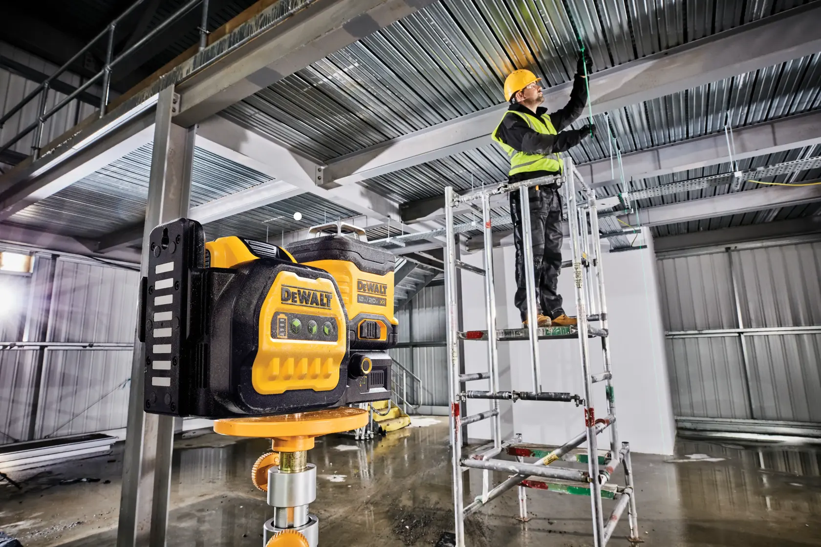 Un nivel láser DEWALT está montado sobre un soporte en un sitio de construcción, mientras un trabajador con equipo de seguridad opera en un andamio al fondo.
