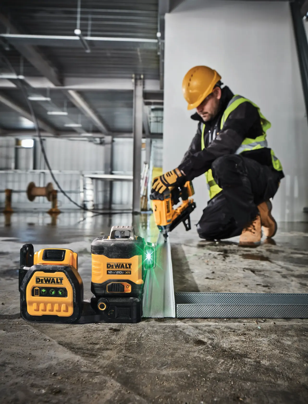 Dispositivo nivelador láser DEWALT y batería colocados en el suelo de una obra, con un trabajador de la construcción usando equipo de seguridad y una herramienta eléctrica al fondo.