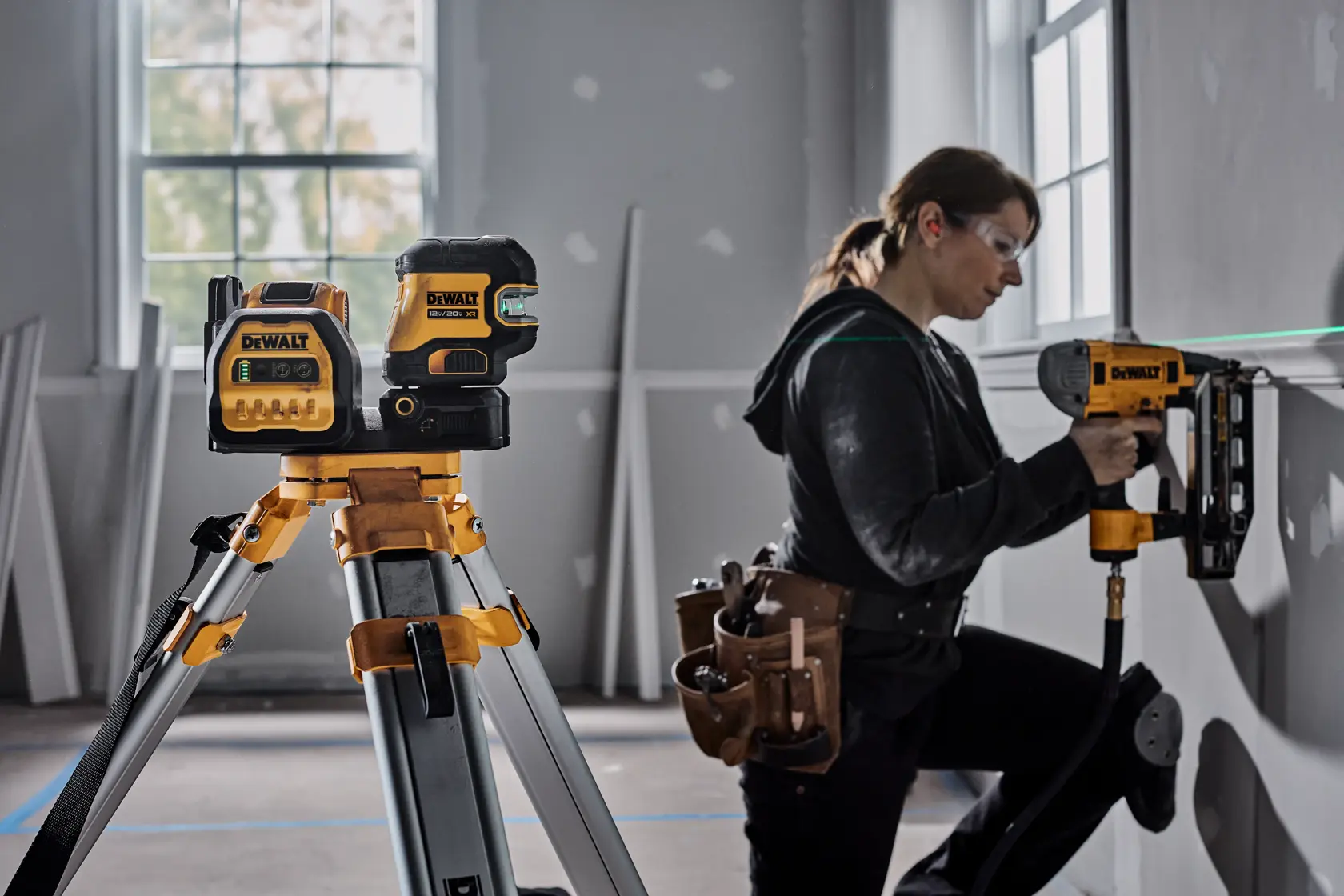 Un nivel láser DEWALT colocado sobre un trípode en una sala de construcción, con una persona usando una herramienta eléctrica DEWALT para trabajar en una pared. El rostro de la persona está difuminado y lleva un cinturón de herramientas.