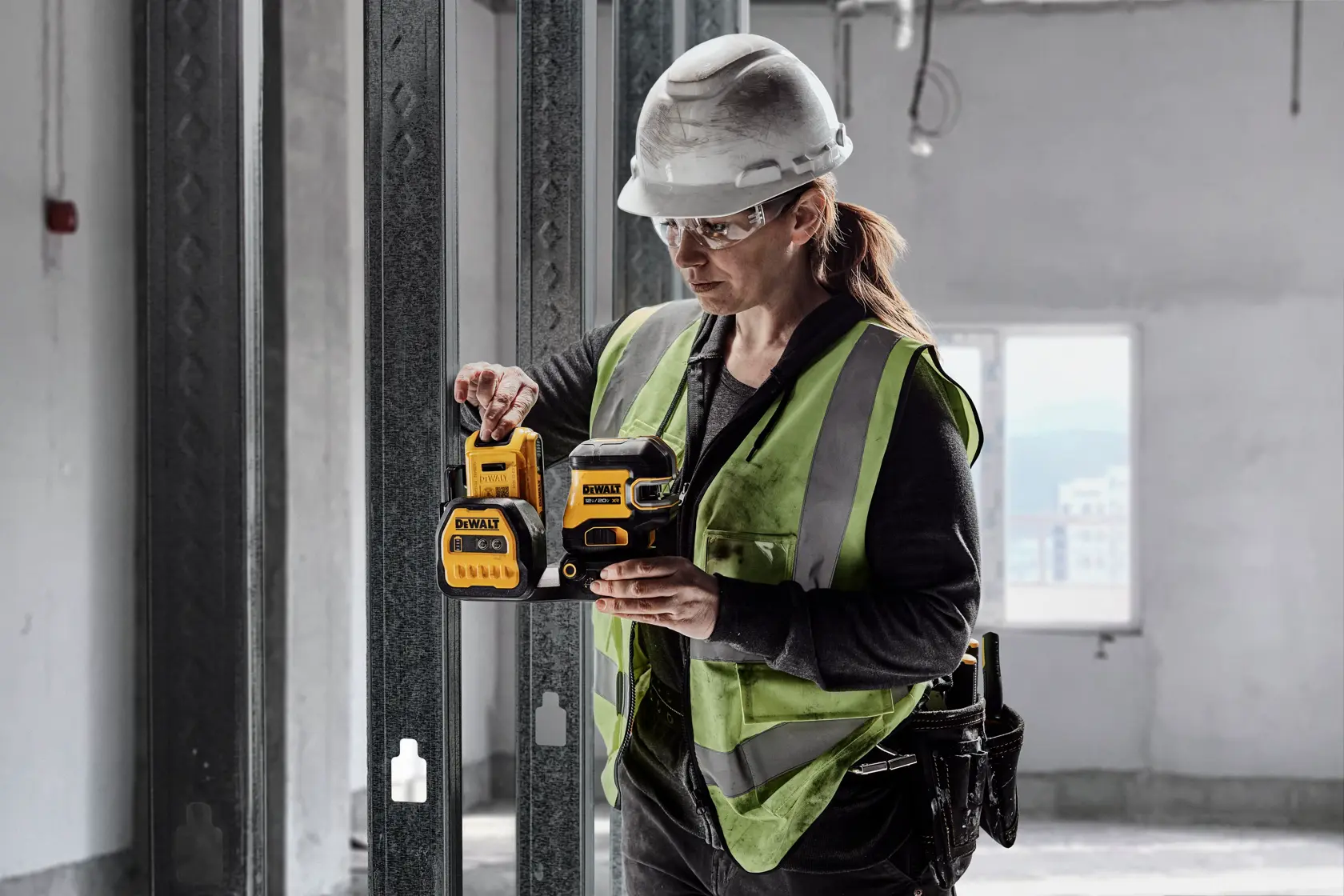 Un trabajador de la construcción con casco y chaleco reflectante utiliza una herramienta niveladora láser DEWALT en una obra. La herramienta es amarilla y negra, y el trabajador está en una habitación sin terminar con montantes metálicos y materiales de construcción.