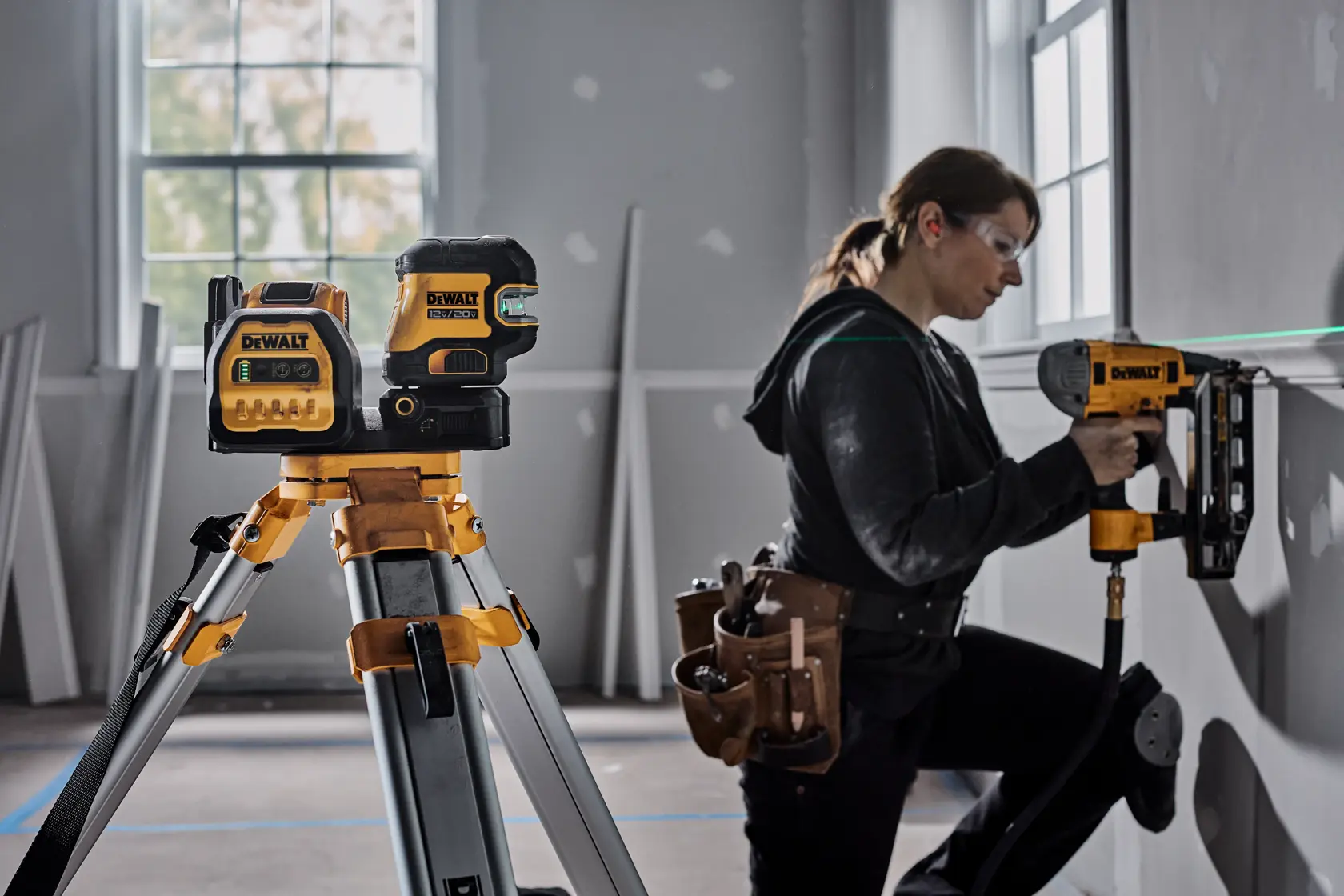 Un nivel láser DEWALT montado en un trípode en primer plano, con un trabajador de la construcción usando una herramienta DEWALT para instalar materiales en una pared en el fondo. El rostro del trabajador está difuminado por privacidad y la escena parece estar dentro de una habitación en remodelación con varios materiales de construcción presentes.