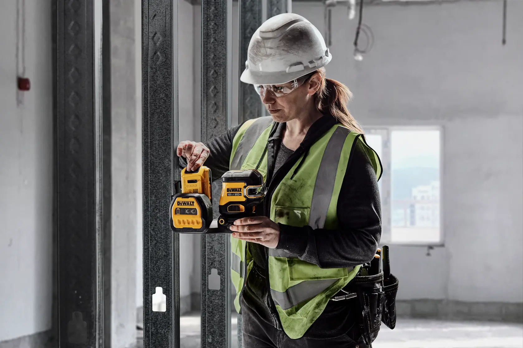 Una persona con chaleco de seguridad y casco utiliza un nivel láser DEWALT en un escenario de construcción. El rostro de la persona está desenfocado y está situada en interiores junto a estructuras metálicas.