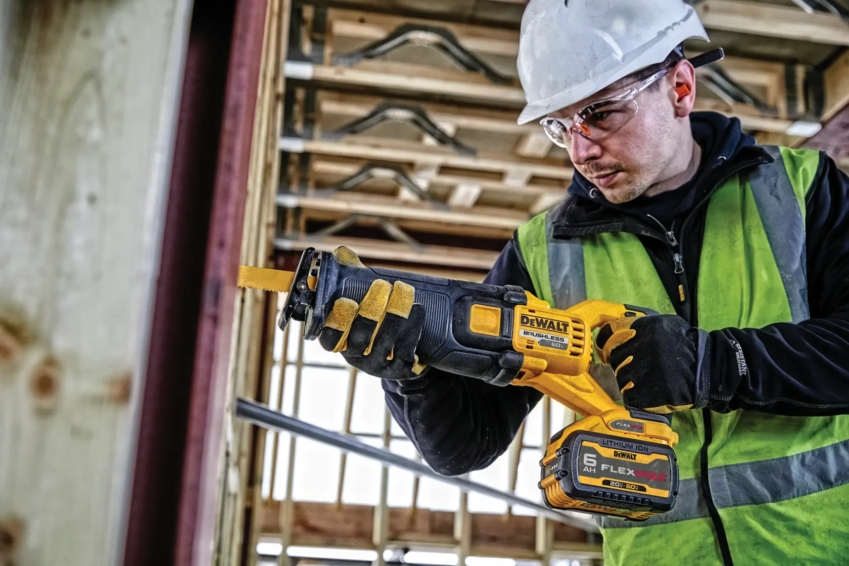 Una persona con casco de seguridad, guantes y chaleco reflectante está usando una sierra recíproca inalámbrica DEWALT amarilla y negra en una obra de construcción.