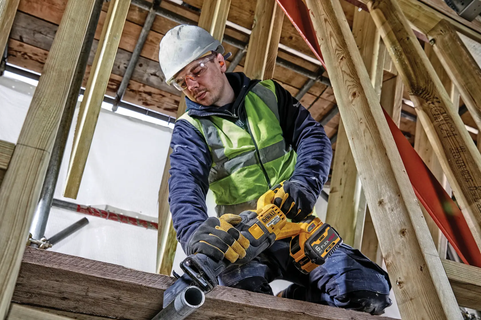 Un trabajador de la construcción con chaleco de seguridad, guantes y casco utiliza una sierra recíproca amarilla inalámbrica DEWALT para cortar una tubería de metal en una obra con vigas de madera.