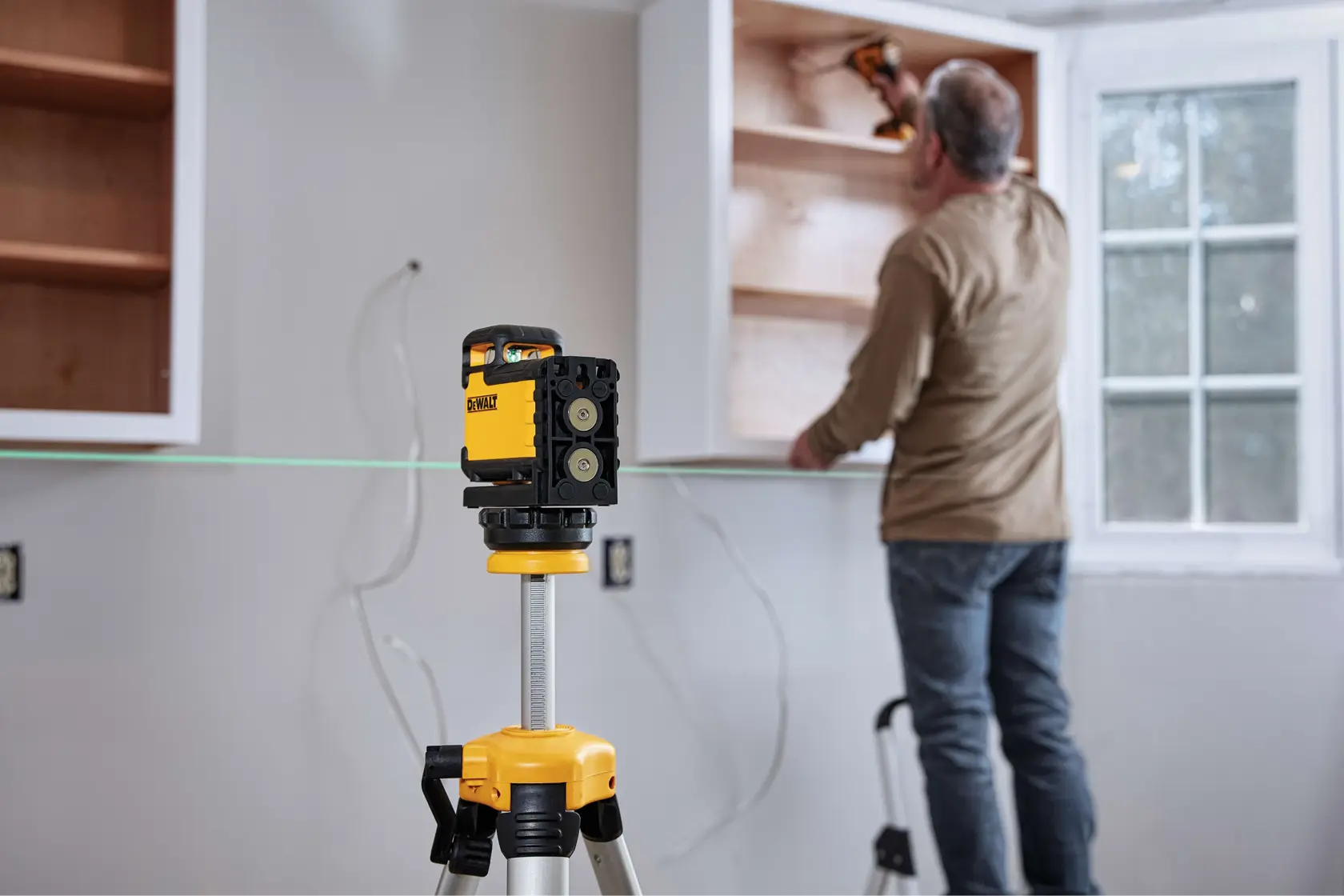 Un nivel láser DEWALT montado en un trípode está posicionado en primer plano mientras una persona instala gabinetes de madera en una pared de una habitación. La línea láser ayuda a asegurar una alineación precisa durante la instalación.