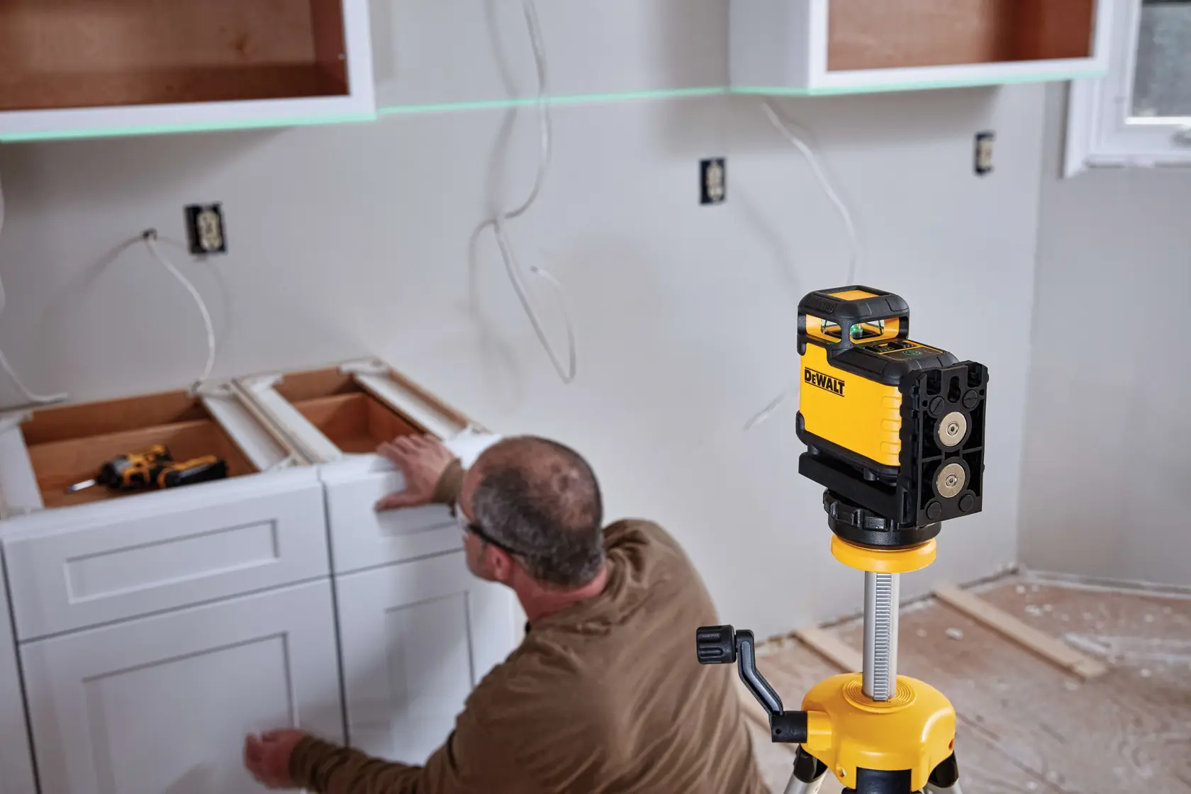 Un nivel láser de línea cruzada verde DEWALT (modelo DW03601CG) montado en un trípode está siendo utilizado en la instalación de una cocina. Una persona está alineando cajones de gabinetes blancos, con herramientas y cableado visibles en la pared y el mostrador.