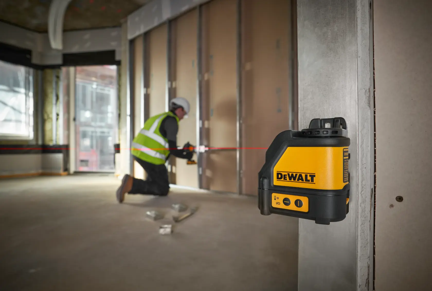 Un nivel láser de líneas cruzadas DEWALT DW088K está montado en un soporte metálico en un sitio de construcción. El dispositivo láser emite líneas rojas para un alineamiento preciso y se pueden ver materiales de construcción en el fondo.