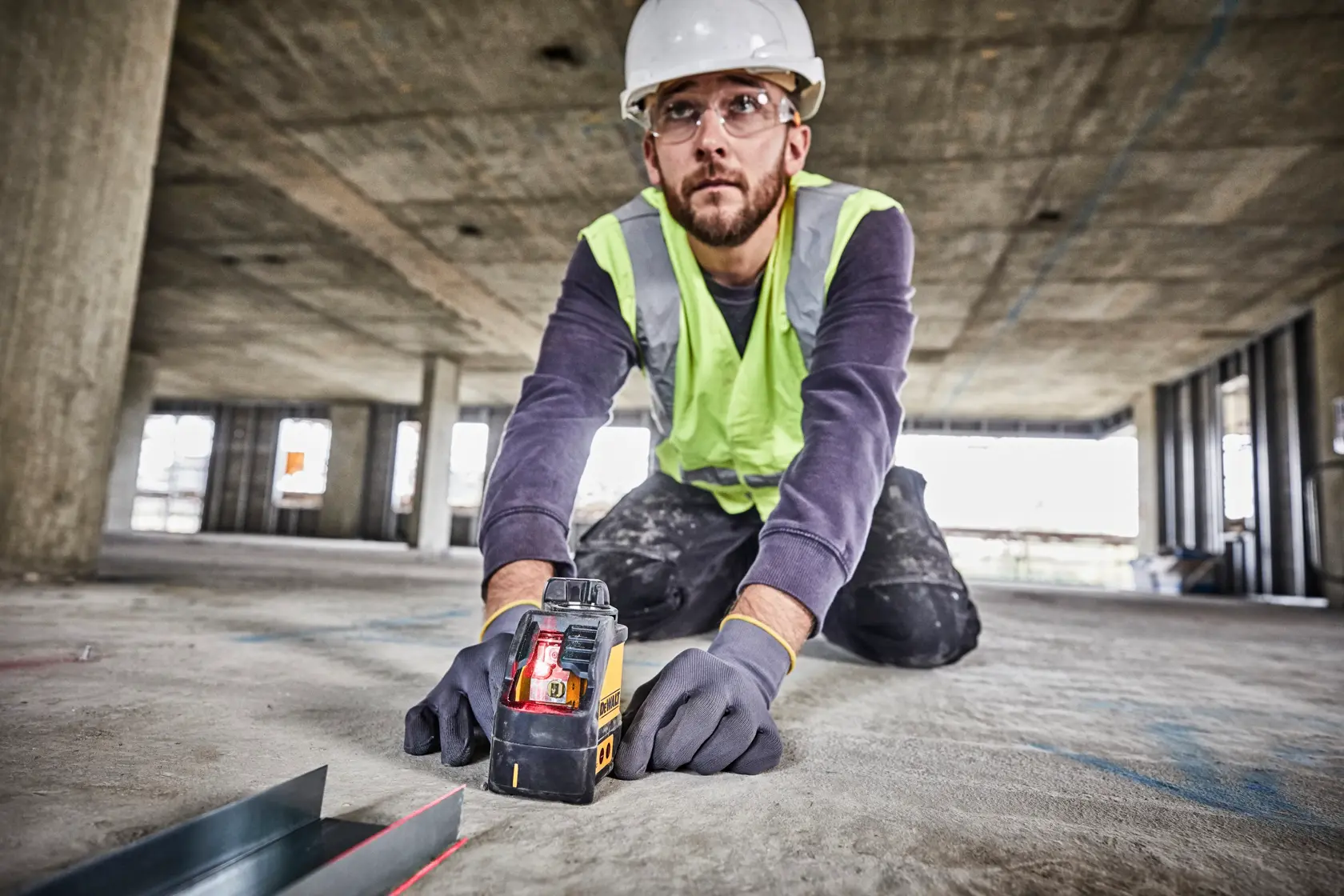 Trabajador usa nivel láser DEWALT DW088K en suelo de obra, con guantes y equipo de seguridad.
