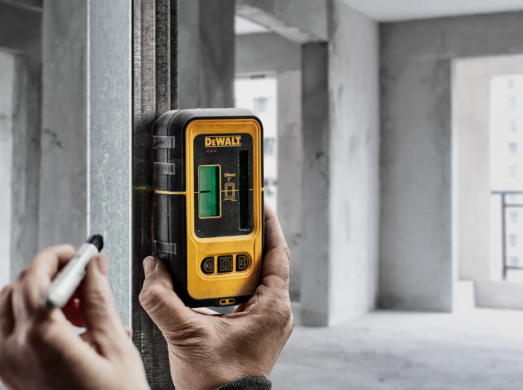 Una persona utiliza un detector de láser verde DEWALT DW0892G fijado a una viga metálica en un sitio de construcción, marcando una línea con un marcador.