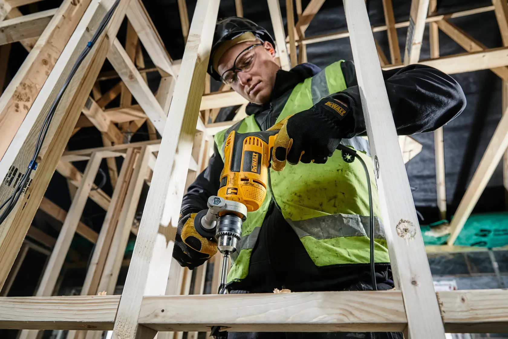 Taladro DEWALT DWD024K usado en vigas de madera en obra por persona con ropa de seguridad.