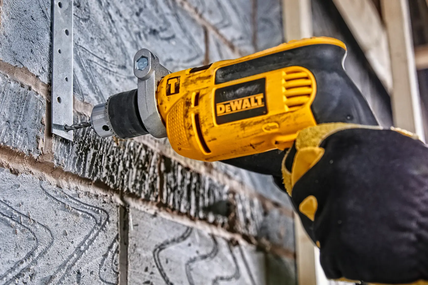 Taladro DEWALT perforando soporte metálico en pared de ladrillo. Mano enguantada sostiene herramienta amarilla.