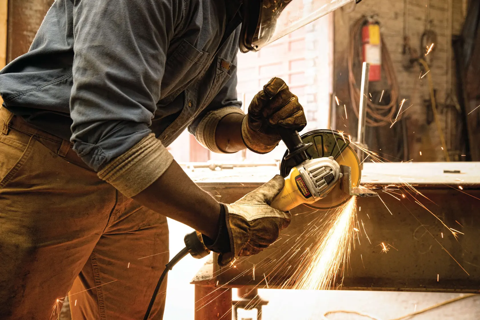 Una persona con guantes protectores y careta utiliza una amoladora angular DEWALT para cortar o pulir metal, con chispas visibles en un taller.