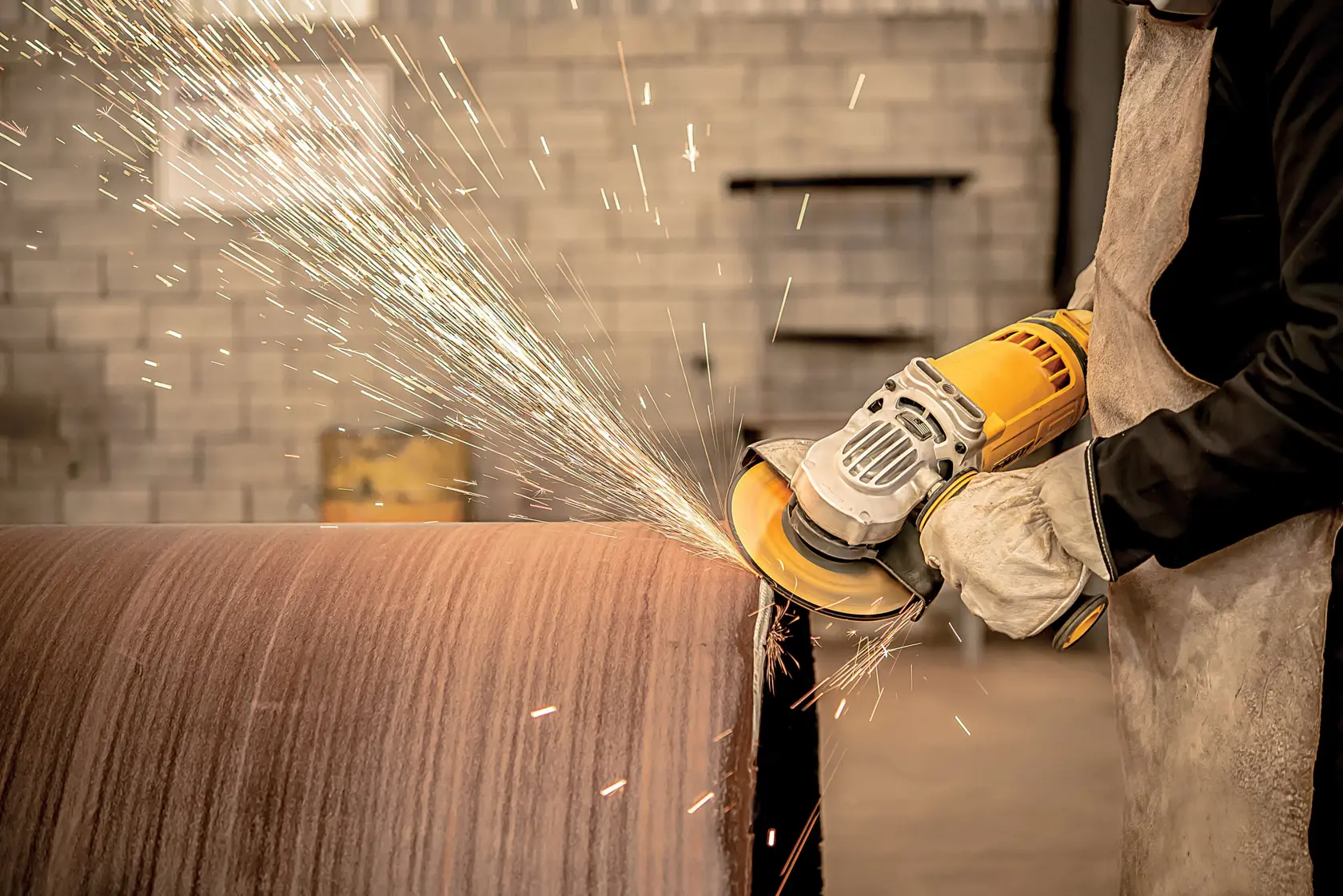 Una persona con guantes y un delantal de protección utiliza una amoladora angular amarilla DEWALT para lijar un gran tubo de metal, generando chispas.
