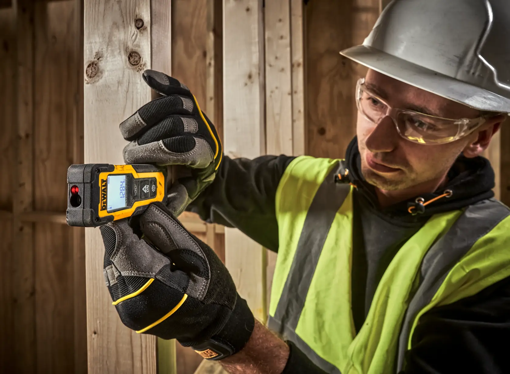 Una persona con chaleco de seguridad, guantes de trabajo y casco está usando un dispositivo medidor de distancia láser DEWALT para medir el ancho de unas tablas de madera en un sitio de construcción. El rostro de la persona está difuminado por privacidad.