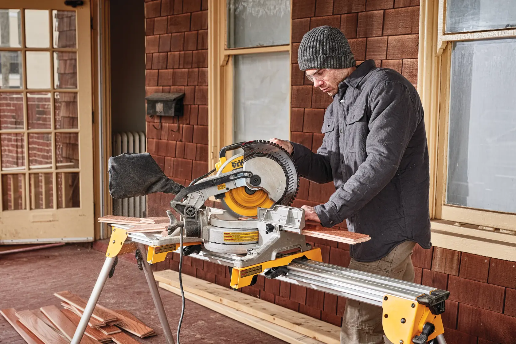 Una persona utiliza una sierra de inglete DEWALT DWS713 para cortar tablas de madera sobre un soporte, llevando una chaqueta y gorro, frente a un edificio con tejas marrones y ventanas.