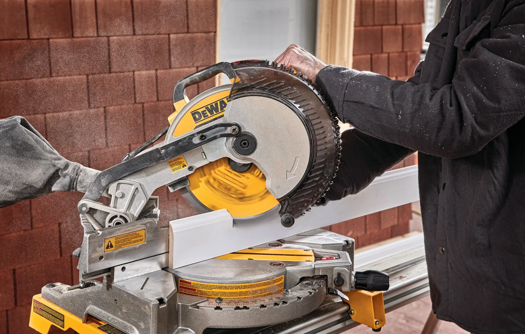 Una persona utiliza una sierra de inglete DEWALT DWS713 para cortar una pieza de moldura blanca. La sierra está montada en un banco de trabajo y se puede ver polvo en el aire.