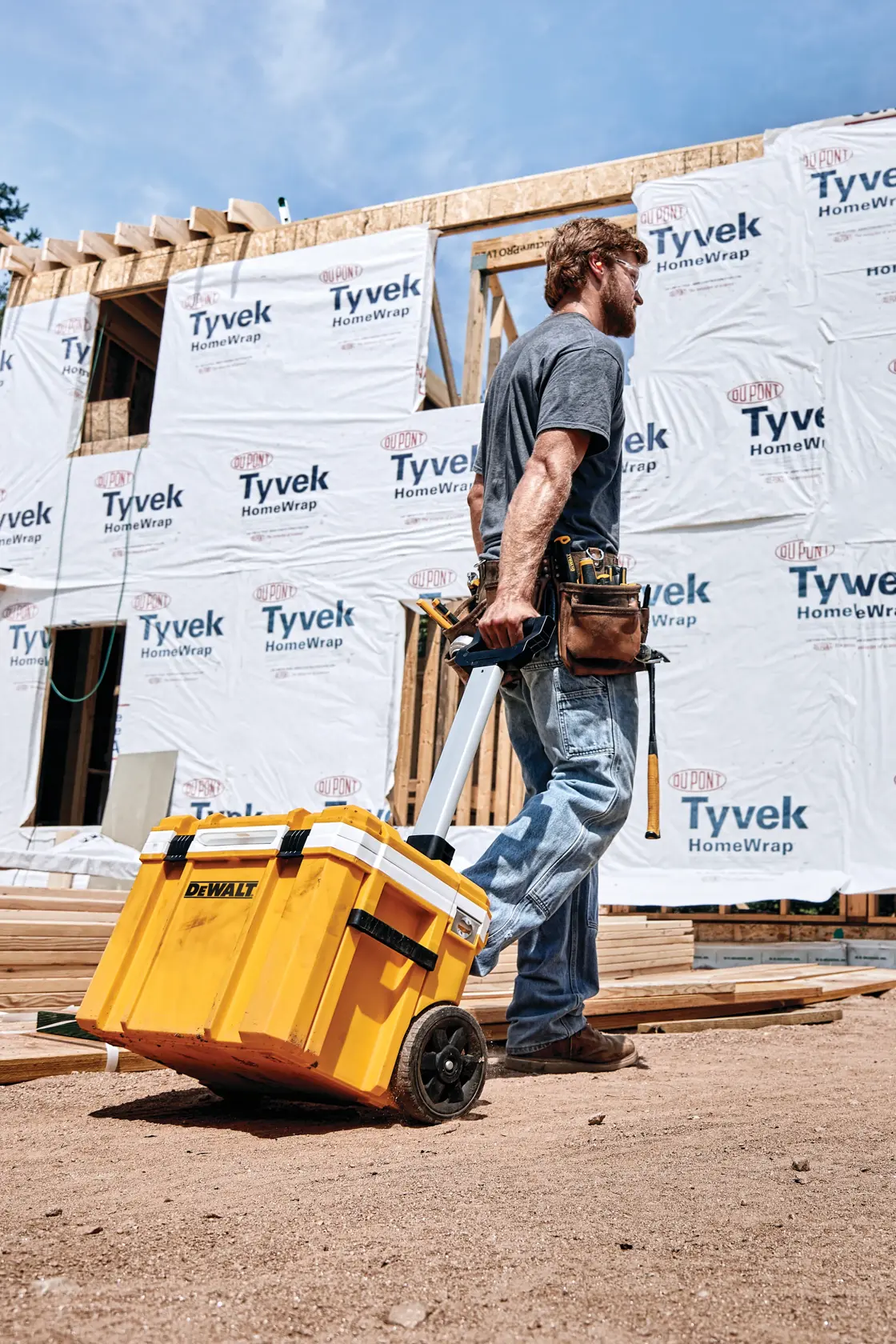 Una persona en un sitio de construcción está tirando de una caja de herramientas amarilla DEWALT con ruedas sobre tierra, frente a un edificio parcialmente envuelto en Tyvek HomeWrap. La persona lleva un cinturón de herramientas y jeans.