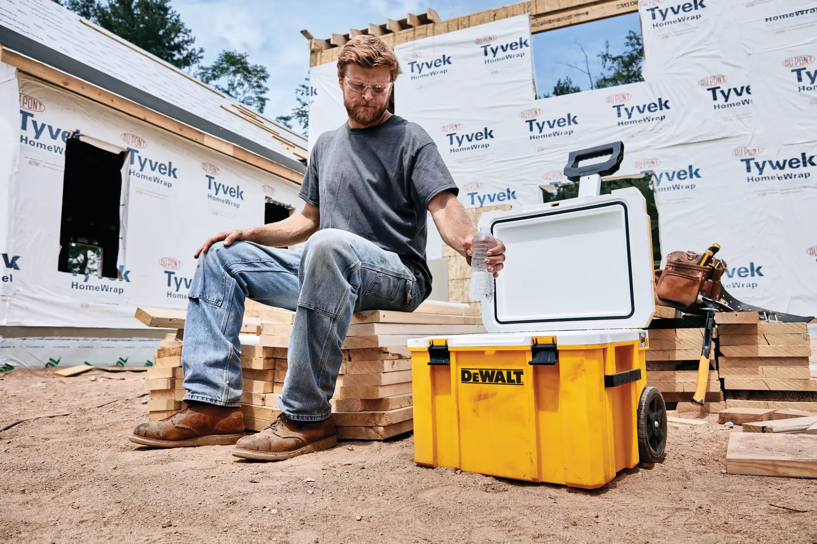 Una persona sentada sobre tablones de madera apilados en un sitio de construcción con una nevera portátil amarilla DEWALT con la tapa abierta a su lado. Se ve una casa parcialmente construida y materiales de construcción en el fondo.