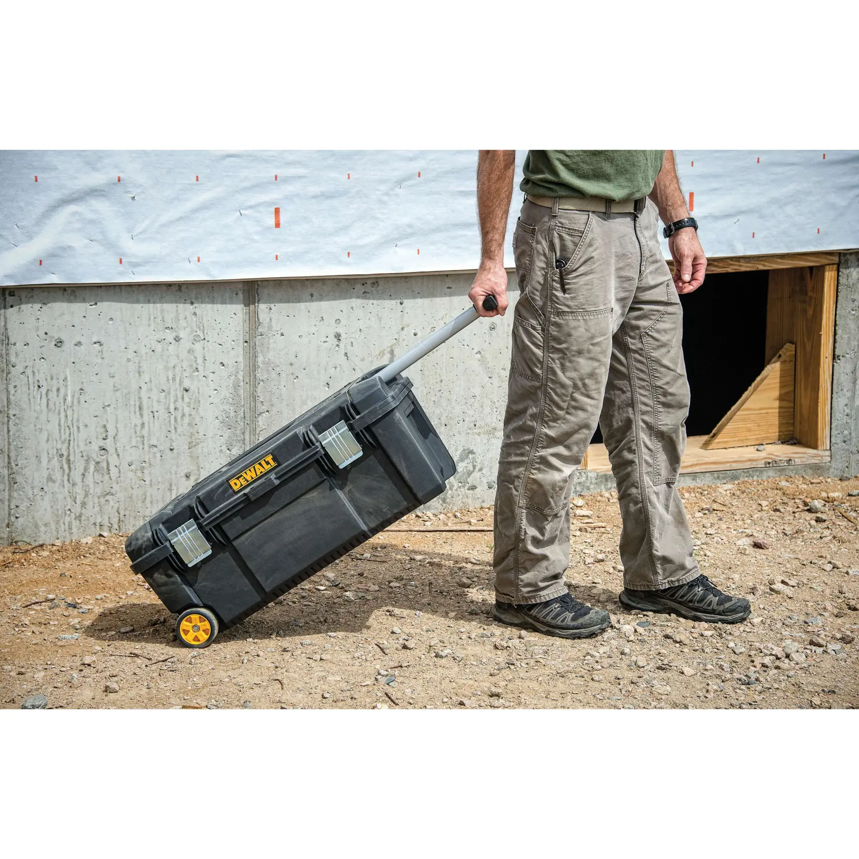 Una persona tira de una caja de herramientas rodante negra DEWALT con ruedas amarillas y un mango telescópico en un sitio de construcción con tierra y una pared de concreto.