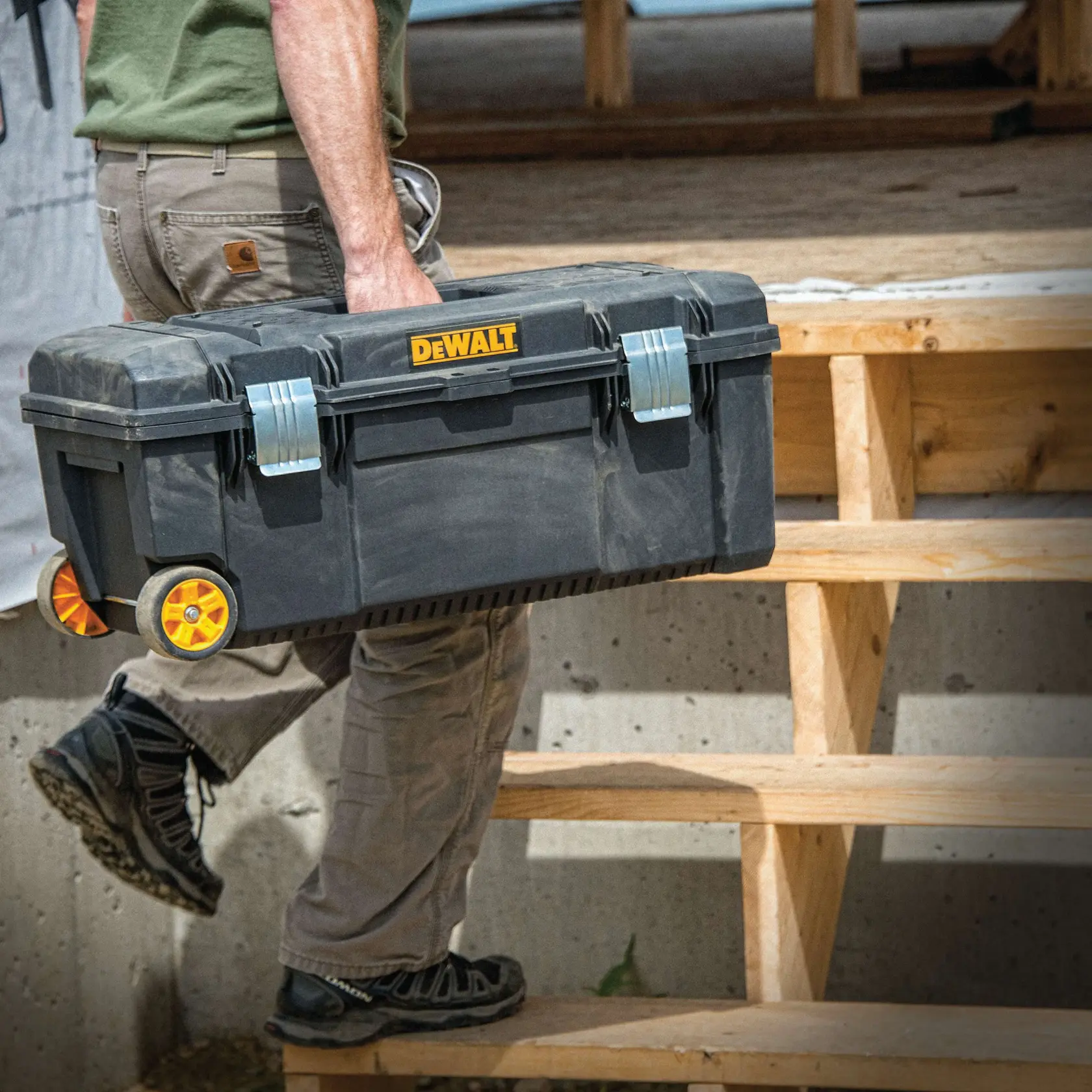 Una persona lleva una caja de herramientas rodante DEWALT subiendo escalones de madera en un sitio de construcción.